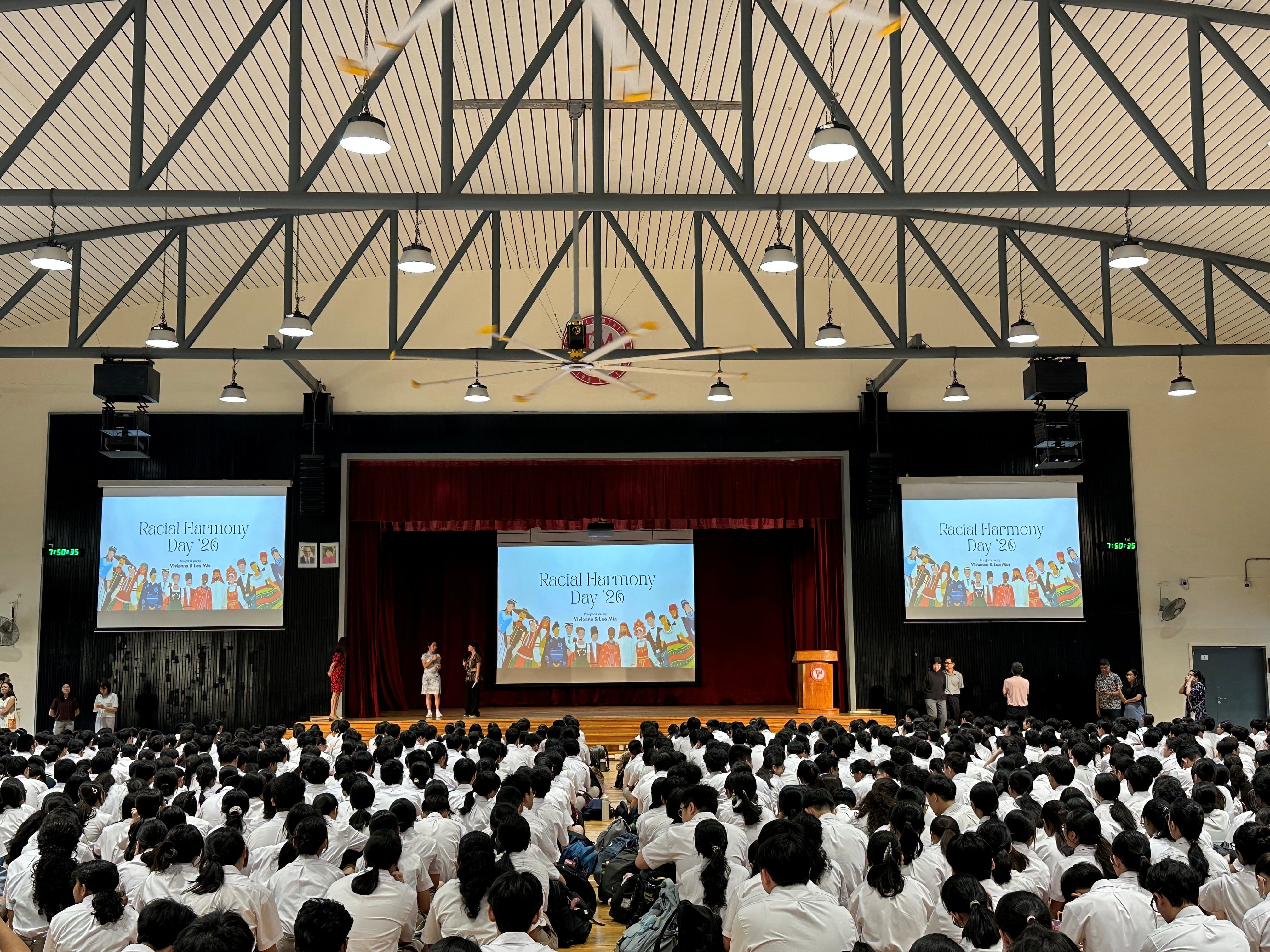 A large school hall packed with students in white uniforms faces a stage, where "Racial Harmony Day '26" is displayed on three screens.